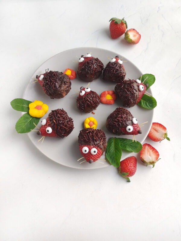 Plate of chocolate-dipped strawberry hedgehogs decorated with candy eyes and mint leaves.