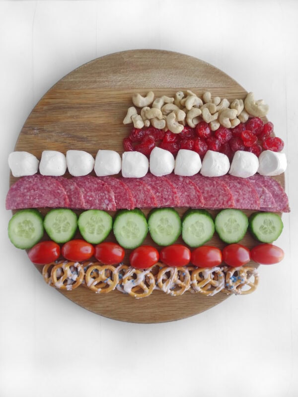 Partially assembled charcuterie board with layered snacks including cucumbers, tomatoes, salami, and marshmallows.
