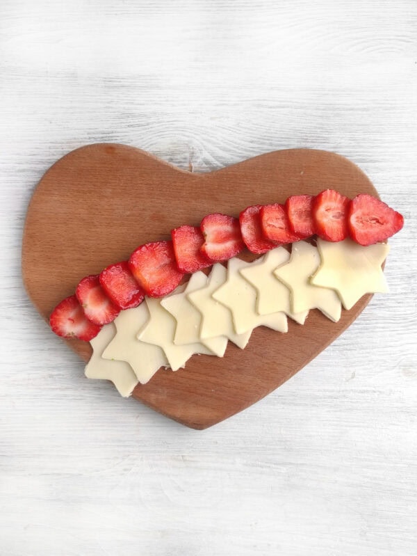 Cheese stars and strawberry slices arranged in alternating rows on a heart-shaped wooden board.