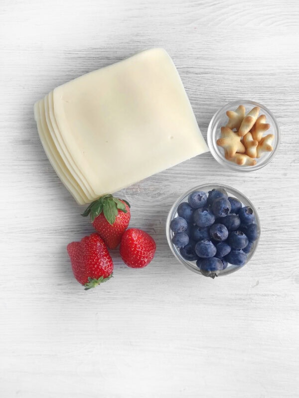 Ingredients for the patriotic charcuterie board including slices of white cheese, strawberries, blueberries, and star-shaped crackers.