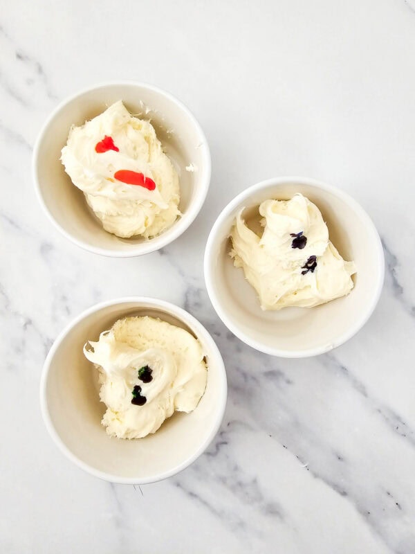 Bowls of white frosting with drops of red, blue, and green food coloring for tinting