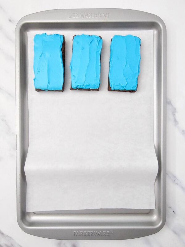 Brownies topped with blue frosting arranged on a baking sheet with parchment paper