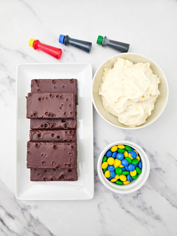 Ingredients for LEGO brownies including brownies, frosting, M&Ms, and food coloring on a white surface