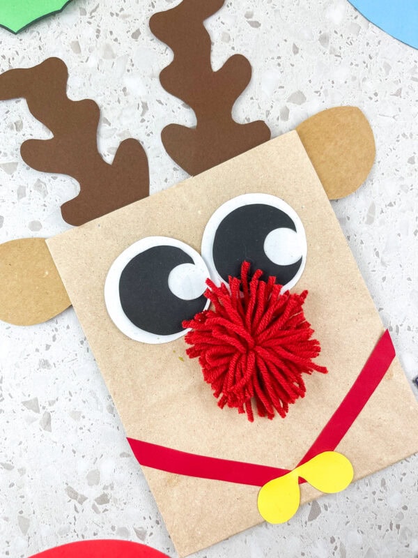 Reindeer paper bag craft with red pom-pom nose, antlers, and yellow bow
