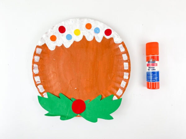 Paper plate gingerbread with glued-on holly bow and glue stick beside it