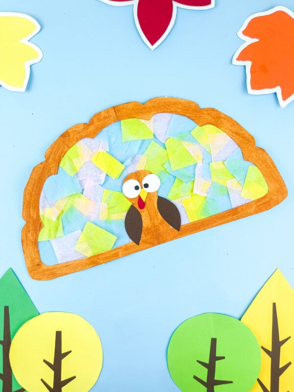 Paper turkey suncatcher craft with colorful tissue paper feathers and cut-out trees.