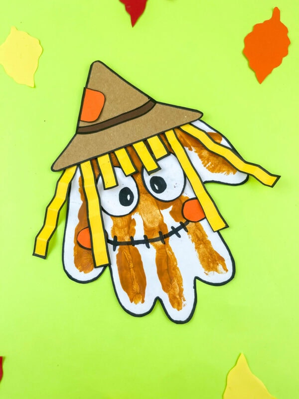 Handprint scarecrow with orange cheeks and a brown hat on green background with leaves