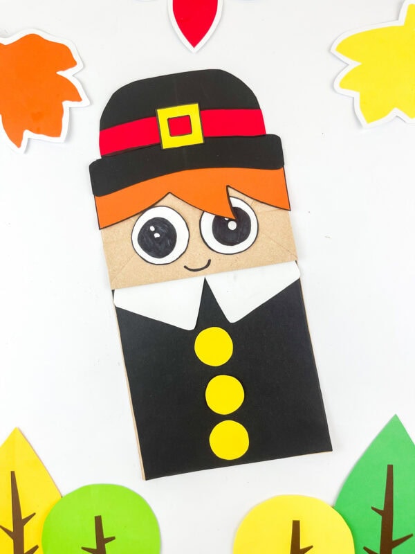 Pilgrim boy paper bag craft with black hat, orange hair, and yellow buttons surrounded by paper leaves.