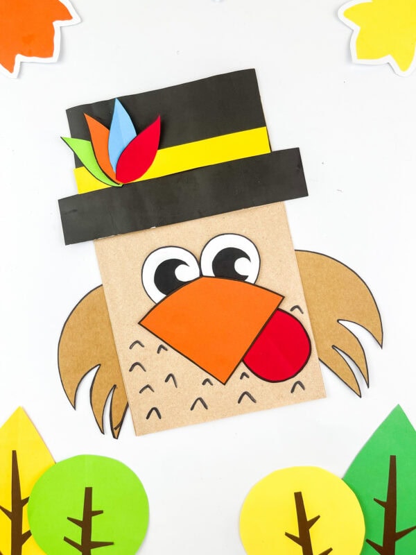 Pilgrim turkey paper bag craft surrounded by colorful autumn trees.