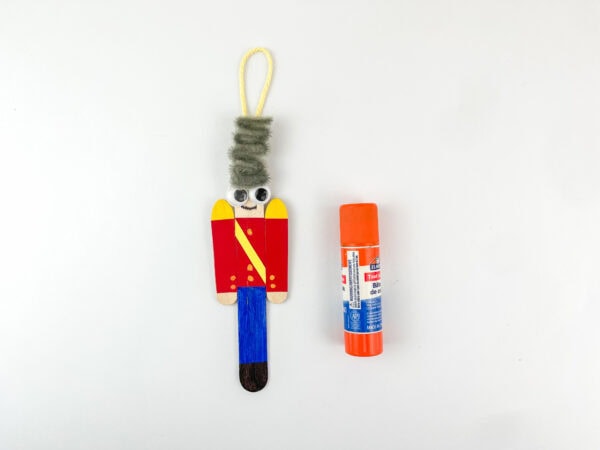 Nutcracker popsicle stick ornament with string loop and glue stick beside it.