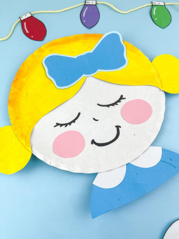 Close-up of the Clara paper plate craft showing smiling face and blue bow under string lights.