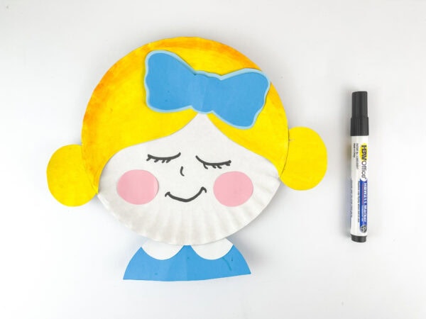 Completed Clara paper plate craft with yellow hair, blue bow, and drawn face beside a black marker.