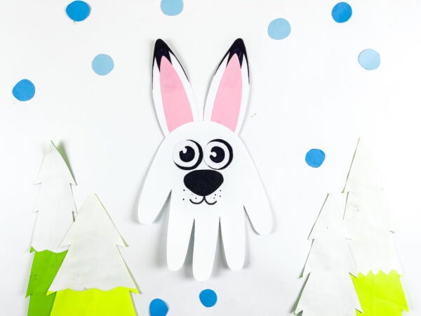 Handprint arctic hare craft placed on a snowy background with trees.