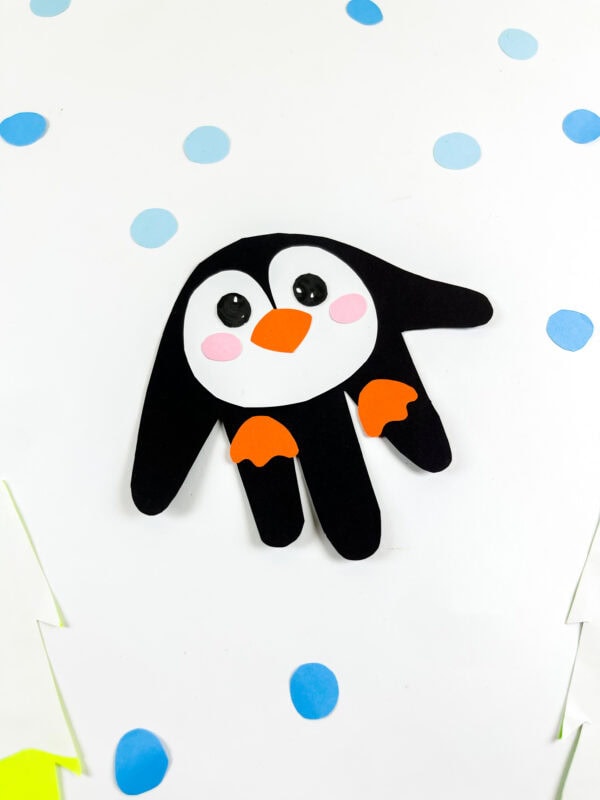 Penguin handprint craft shown on a snowy background with scattered blue dots.