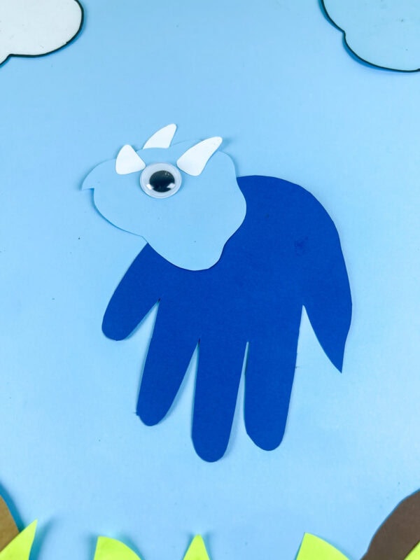 Blue triceratops handprint craft on sky background with grass