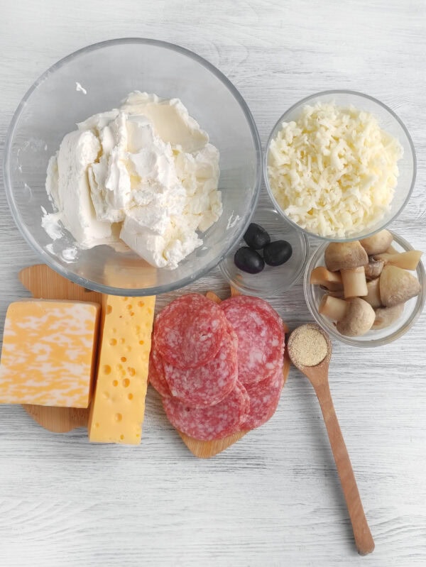 Ingredients for hedgehog cheese spread including cream cheese, shredded cheese, salami, olives, mushrooms, and seasoning