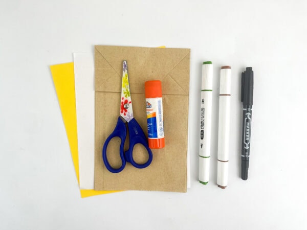 Craft supplies including a brown paper bag, colored paper sheets, scissors, a glue stick, and markers on a white surface.