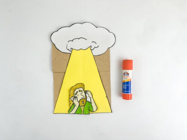 The Saul character cutout glued onto the yellow light beam on the paper bag, next to a glue stick.