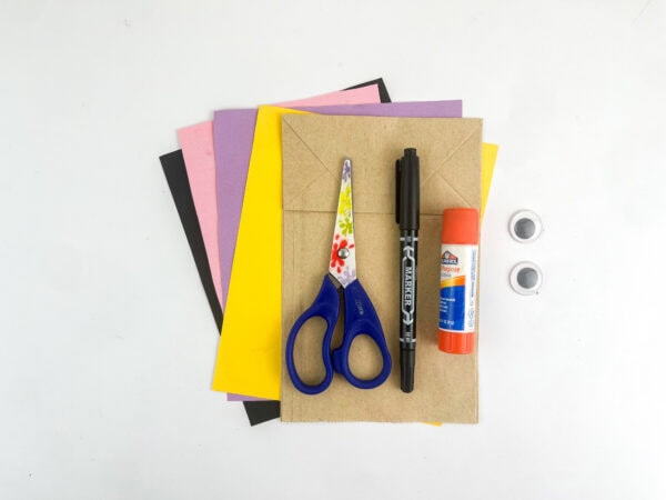 Craft supplies laid out: colored paper, a paper bag, scissors, marker, glue stick, and googly eyes