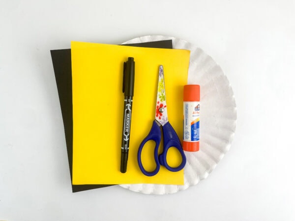 Craft supplies including a white paper plate, yellow and black construction paper, a black marker, scissors, and a glue stick on a white surface.
