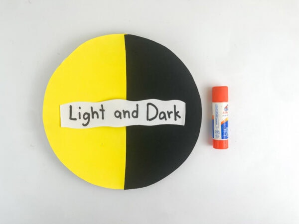The handwritten "Light and Dark" paper strip placed across the center of the yellow and black paper circle, next to a glue stick.