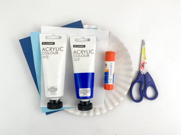 Craft supplies arranged on a white background including construction paper, a white paper plate, two tubes of acrylic paint, a glue stick, and scissors.