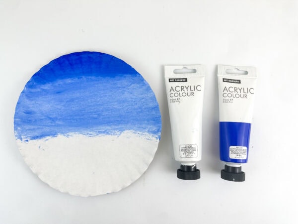 A paper plate painted with a blue gradient next to two tubes of white and blue acrylic paint.