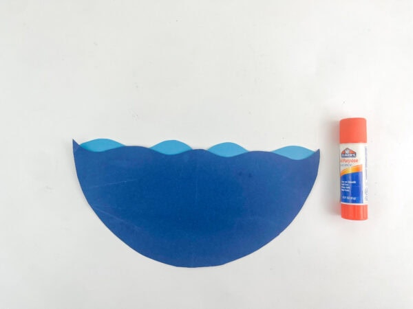 A light blue wavy paper strip glued behind a larger dark blue wavy paper cutout next to an orange glue stick.