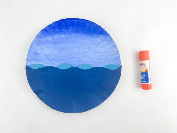 Wavy blue paper water layers glued onto the bottom half of a gradient-painted blue paper plate next to an orange glue stick.