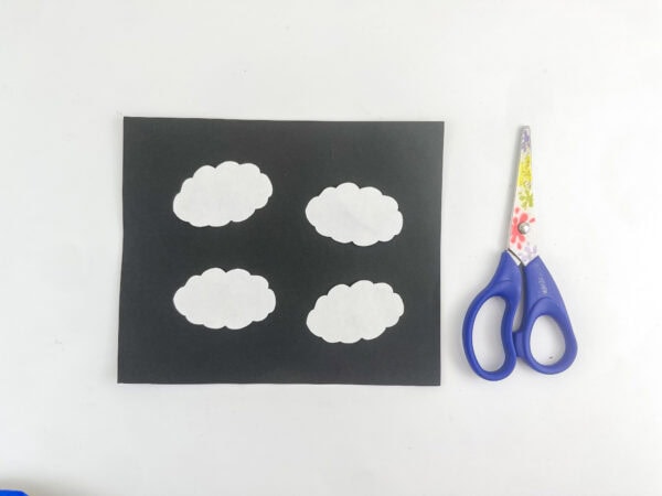 Four white paper cloud cutouts on a piece of black paper next to a pair of scissors.