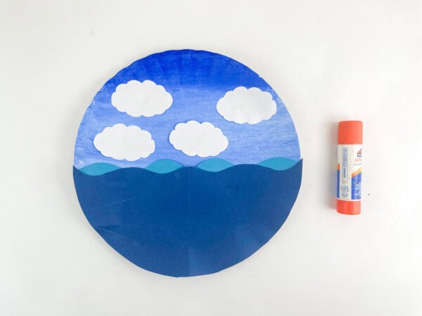 Four white paper clouds glued onto the sky portion of the blue paper plate craft next to an orange glue stick.