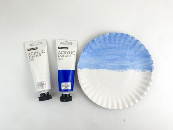 A paper plate with its top half painted light blue, positioned next to white and blue tubes of acrylic paint.