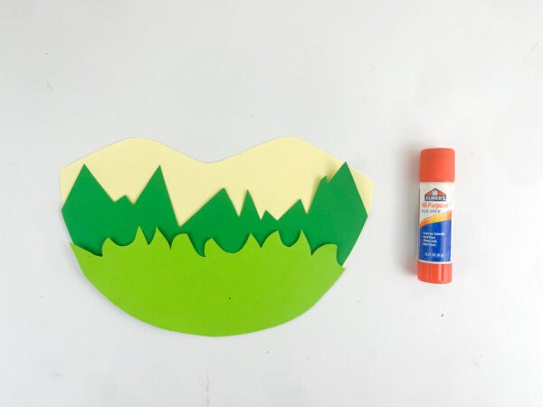 Three layers of cut construction paper glued together to form a landscape background next to an orange glue stick.