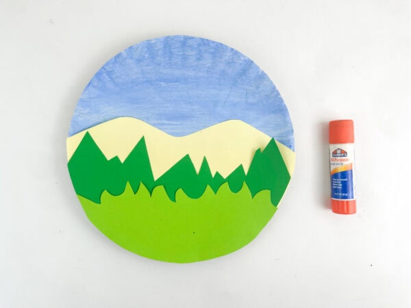 The assembled construction paper landscape background glued onto the lower half of the blue-painted paper plate next to a glue stick.