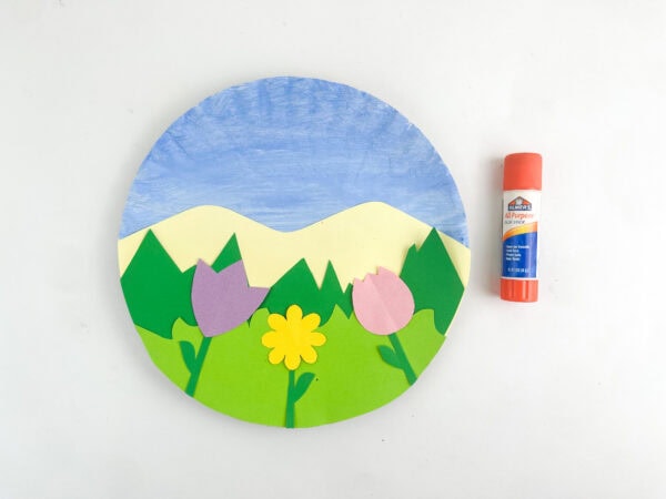 Three paper flowers glued onto the paper plate landscape scene next to a glue stick.