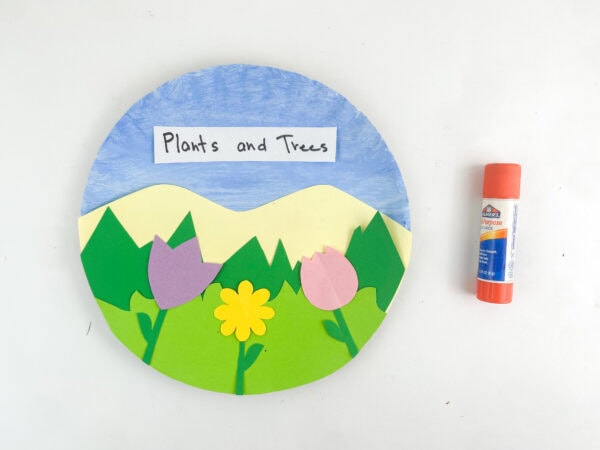 Completed paper plate craft labeled "Plants and Trees" sitting next to an orange glue stick on a white surface.