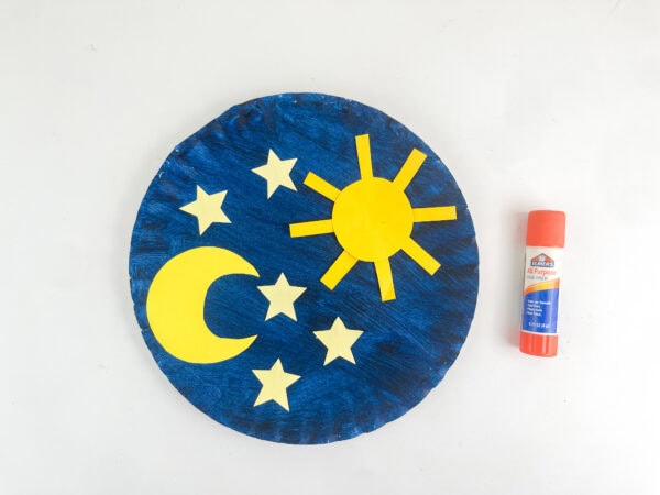 Yellow paper sun, moon, and stars glued onto the blue paper plate next to a glue stick.