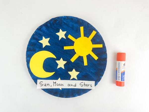 The finished paper plate craft with the "Sun, Moon and Stars" label attached, next to a glue stick.