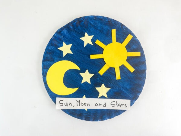 A completed blue paper plate craft with a yellow paper sun, crescent moon, stars, and a handwritten "Sun, Moon and Stars" label on a white background.