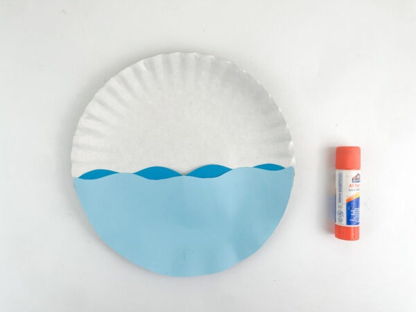 Blue paper waves glued onto the bottom half of a white paper plate next to a glue stick.