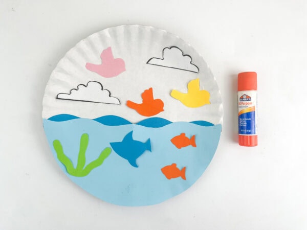The paper plate craft with bird, cloud, fish, and seaweed cutouts glued in place next to a glue stick.