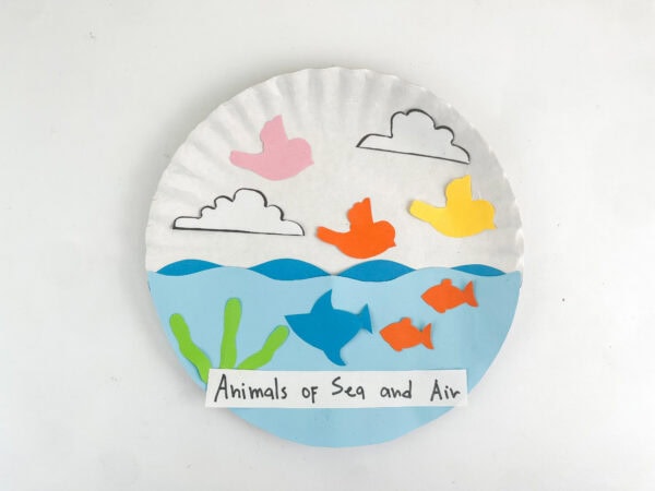 A completed paper plate craft on a plain white background showing paper cutouts of birds, clouds, fish, waves, and a handwritten label reading "Animals of Sea and Air".