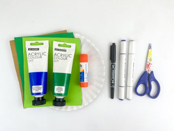 Craft supplies including acrylic paint tubes, colored paper, a glue stick, a white paper plate, markers, and scissors.