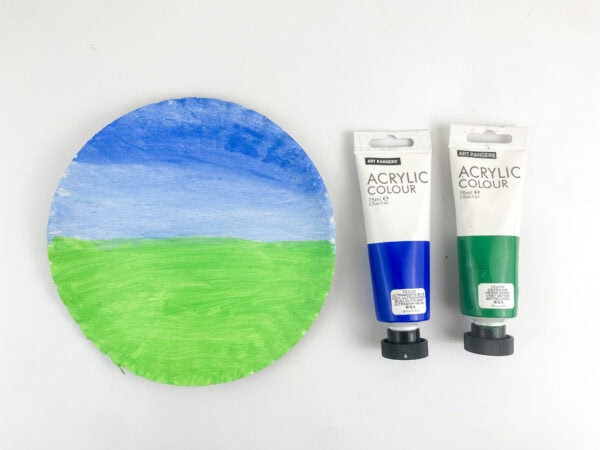 A paper plate painted blue and green next to two tubes of blue and green acrylic paint.