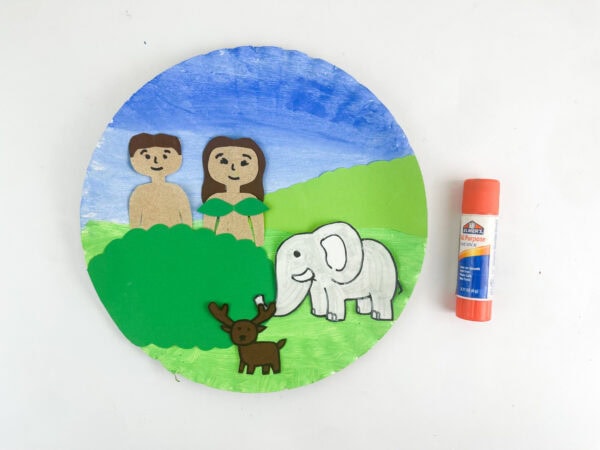 The assembled paper plate craft with the figures and landscape glued down, shown next to an orange glue stick.