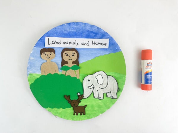 An assembled paper plate craft depicting Adam, Eve, a deer, and an elephant with an orange glue stick next to it.