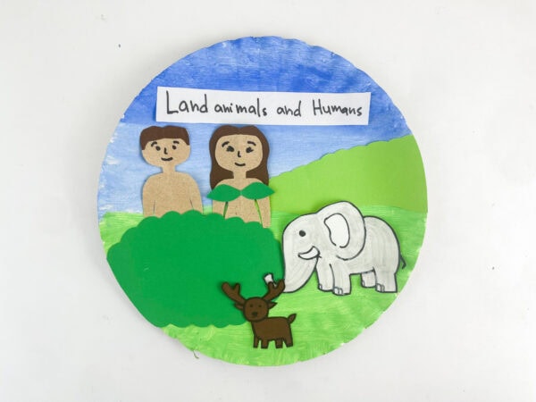 A completed paper plate craft showing human and animal figures on a painted blue and green landscape background.