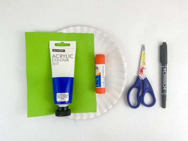 Craft supplies arranged on a white surface, including blue paint, green paper, a paper plate, a glue stick, scissors, and a black marker.