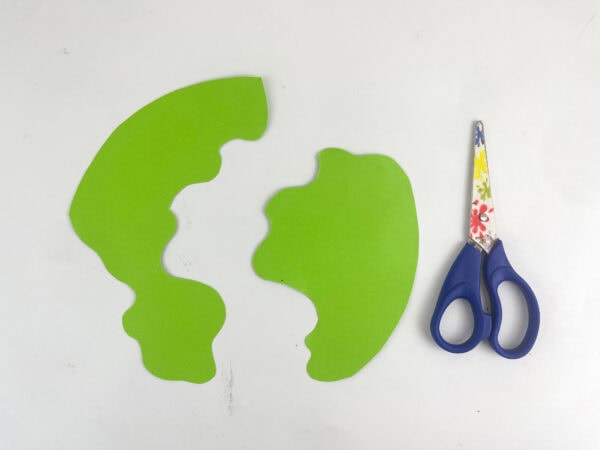 Two green paper cutouts resembling continents sitting next to a pair of blue scissors.