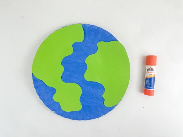 Green paper continents glued onto a blue painted paper plate, with an orange glue stick next to it.